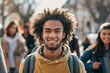 Radiant young man with curly hair smiling broadly, wearing a yellow sweatshirt and backpack, enjoying a sunny day outside with blurred friends in the background, representing youth and happiness