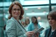 © Enigma - Professional woman in a light grey blazer smiles confidently at the camera, holding a digital tablet with colleagues blurred in the background within a modern office setting