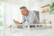 Contented mature man with a smartwatch browsing smartphone by a glass-top counter in a sunny kitchen setting