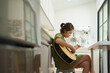 Focused Asian woman playing acoustic guitar in a modern kitchen, sitting on a stool