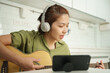 Focused Asian woman learning guitar with online tutorial, headphones on, in a white-tiled kitchen