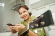 Cheerful young Asian woman with headphones playing guitar and using smartphone in a modern kitchen
