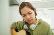 Intently practicing guitar, young Asian woman in headphones at home, focused on chords in a kitchen setting