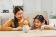 Cute little asian girl saving money by adding a coin in piggy bank with mother at home