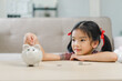 Cute little asian girl saving money by adding a coin in piggy bank with mother at home