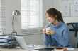 Smiling young woman enjoying a warm beverage during a break in a modern home office. Work life balance concept