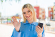 Young pretty blonde woman using mobile phone at outdoors saluting with hand with happy expression