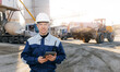 Modern technologies industrial cement plant concept. Engineer in uniform and hard hat uses tablet computer to control equipment trucks at production site