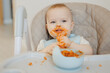 Happy messy baby boy eating pasta with tomato enjoys food sits In high chair, light background