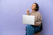 © luismolinero - Young mixed race woman sitting on a chair with laptop isolated on purple background pointing up and surprised