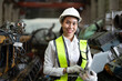 Confident Asian female engineer with labtop computer smiles while wearing a white hard hat and reflective vest in a mechanical workshop.