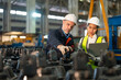 Senior male engineer and Asian female engineer collaborate using a laptop amidst heavy machinery in an industrial factory setting.