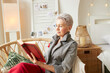 Side view of elegant stylish aged female with short haircut in gray denim jacket reading fascinating novel relaxing in wicker chair among cozy interior decor spending weekend in country house