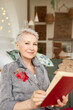 Pretty charming aged caucasian woman grandma with stylish short hair with cute smile reading novel out loud looking at camera and holding hardcover book, dressed in denim jacket with red brooch