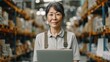 © iuricazac - Asian woman in warehouse smiling holding laptop wearing apron standing in front of shelves with boxes.