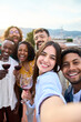 © CarlosBarquero - Group of diverse friends taking selfie and having fun at rooftop home party holding glass of red wine. Beautiful young people. Smiling community drinking on outdoor terrace. Positive friendship