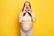 Screaming excited young Caucasian pregnant woman dressed in top posing against bright yellow wall keeps hands near her mouth making announcement yelling loud
