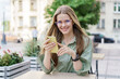 © muse studio - A cheerful young woman in glasses uses her smartphone at an outdoor cafe, embodying a relaxed and connected lifestyle.