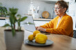 Senior happy Caucasian woman sitting in kitchen at home and using laptop to pay bills