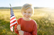 Cute little boy celebrating of July, 4 Independence Day of USA at sunny summer sunset. Child running with american flag of United States on wheat field.