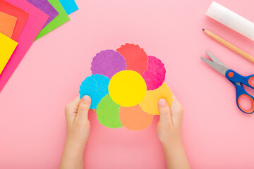 Wall Mural - Baby girl hands holding and showing beautiful flower head shape from colorful paper on pastel pink table background. Closeup. Point of view shot. Little child creating decoration. Top down view.