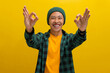 Smiling young Asian man, dressed in a beanie hat and casual shirt, is gesturing the OK sign with his hand while looking excitedly at the camera. He stands against a yellow background