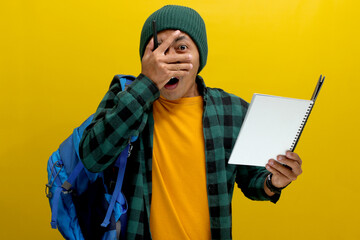 Funny Asian student, wearing a beanie hat and casual clothes, carrying a backpack, appears shocked while reading his notebook, seemingly forgetting his homework, and preparation for an upcoming exam