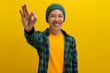 Smiling young Asian man, dressed in a beanie hat and casual shirt, is gesturing the OK sign with his hand while looking excitedly at the camera. He stands against a yellow background