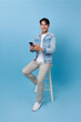 Portrait happy smiling young asian man using smartphone sitting on chair isolated on blue background. People And Technology Concept.