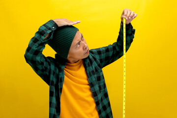 An Asian man, dressed in a beanie hat and casual clothes, measures his height using a yellow measuring tape while standing against a yellow background