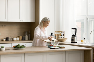 Pretty blonde senior woman chopping fresh vegetables on board on kitchen table, throwing slices into bowl, preparing healthy meal for dinner, cooking salad from organic food ingredients