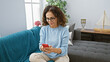 © Krakenimages.com - Middle-aged hispanic woman with curly hair using smartphone in cozy living room interior.
