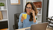 © Krakenimages.com - A professional middle-aged hispanic woman with curly hair multitasks in an office, talking on the phone and holding a coffee mug.