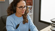 © Krakenimages.com - A mature hispanic woman with curly hair wearing glasses and a headset focuses at work in a modern office interior.