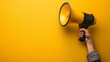 © P - A hand is shown holding a megaphone against an yellow background, symbolizing communication or announcement. The image is dynamic and attention-grabbing, with copy space