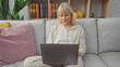 © Krakenimages.com - A blonde woman works on a laptop seated on a gray sofa indoors, exuding a casual and focused ambiance.