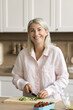 © fizkes - Cheerful pretty mature retired woman preparing salad for dinner in home kitchen, looking at camera with toothy smile, cutting cucumber, slicing fresh vegetables on chopping board