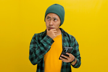 Wall Mural - A puzzled and confused Asian man, dressed in a beanie hat and casual shirt, reacts to news on his mobile phone, displaying confusion, unhappiness, and deep contemplation, isolated on yellow background