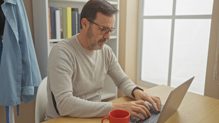 Sticker - A mature man with a beard working on a laptop in a bright home office space.