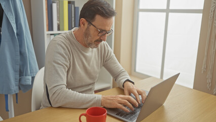 Poster - Mature bearded man typing on laptop at home office with glasses and red mug.