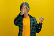 Excited young Asian man, dressed in a beanie hat and casual shirt, samples food with a spoon, giving an OK hand gesture, smiling at the camera, expressing satisfaction with the taste yellow background
