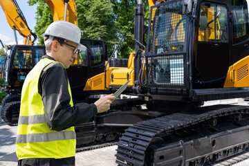 Aufkleber - Engineer in a helmet with a digital tablet stands next to construction excavators..