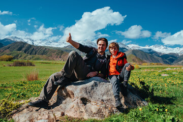 Poster - Happy father and son hikers sitting on rock in the mountains and showing thumbs up