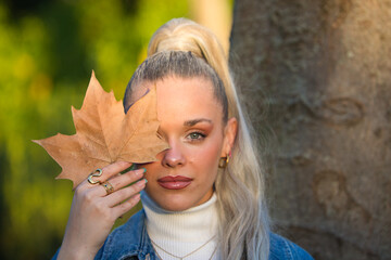 Pretty young blonde woman with a ponytail in her hair covers one eye with a dry leaf. Focus on the leaf.