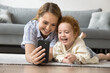 Cheerful young mom and cute curly haired toddler boy taking selfie on mobile phone at home, resting on floor, using online Internet learning educating application
