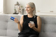 Positive satisfied senior bank customer woman sitting on home couch with blue credit card and smartphone, looking away with dreamy face, thinking, planning shopping on Internet