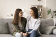 Positive mom and teenager kid enjoying conversation, friendship, leisure at home, holding hands, sitting on sofa close, talking, smiling, laughing, celebrating happy girls success