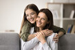 Happy lovely teenage girl in glasses hugging beautiful mom from behind with love, trust, gratitude, looking at camera with healthy toothy smile. Cheerful mother and teen kid home family portrait