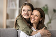 Happy teenage daughter kid in stylish glasses hugging pretty mom from behind, looking away with thoughtful dreamy face, smiling. Mother and teenager relaxing at home, thinking, planning family leisure