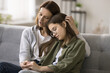 Compassionate young mother comforting depressed sad teen girl, hugging daughter with love, care, speaking, giving sympathy, advice, support, holding kids hand, sitting on home couch